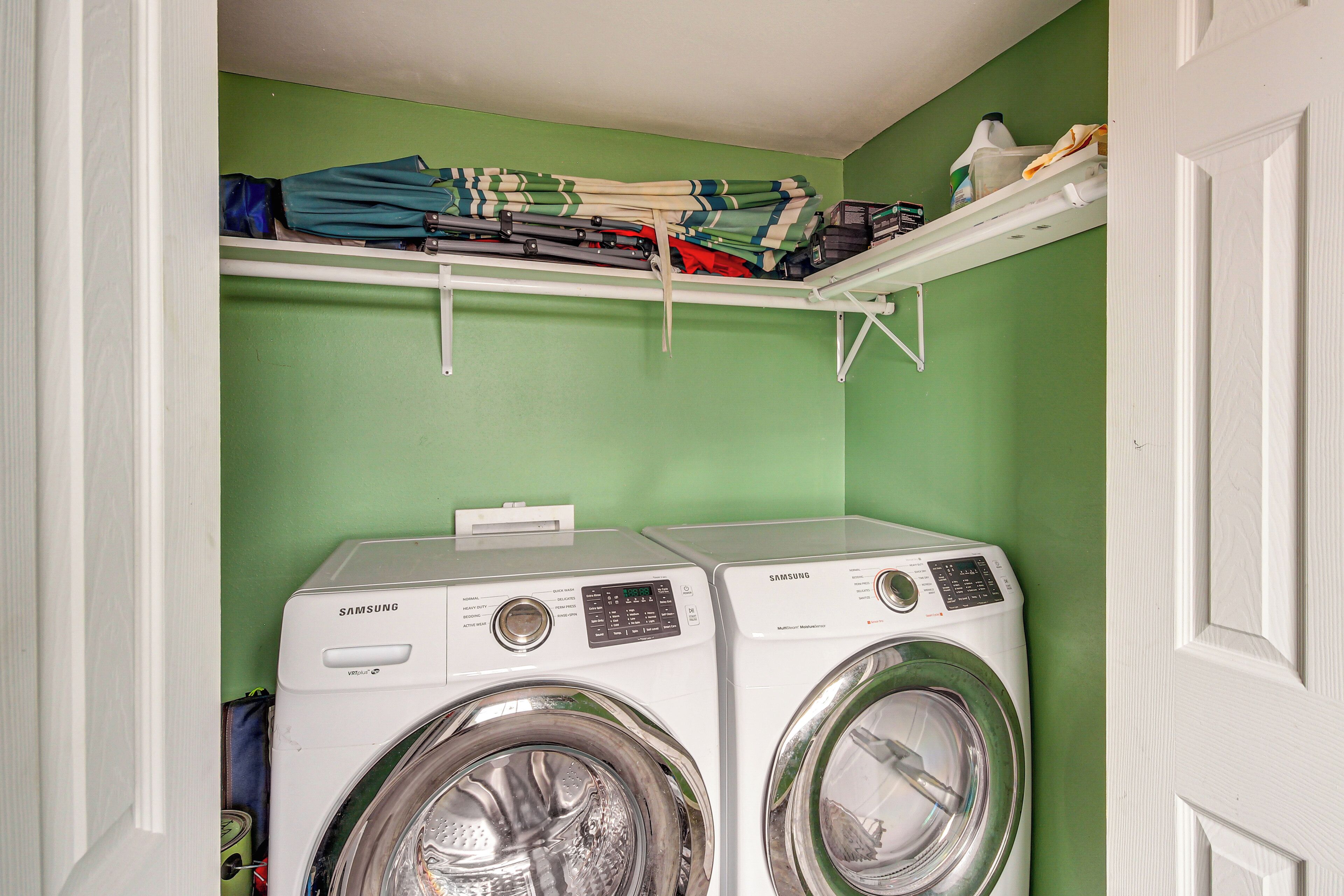 A laundry room with two Samsung washing machines, a shelf above with folded clothes, and a green wall.