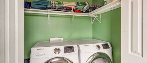 A laundry room with two Samsung washing machines, a shelf above with folded clothes, and a green wall.