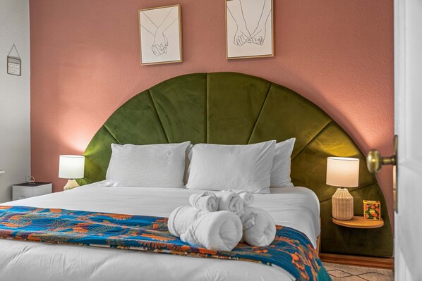 Une chambre d’hôtel avec un grand lit, deux lampes de chevet, une tête de lit recouverte d’un tissu vert et un plaid coloré.