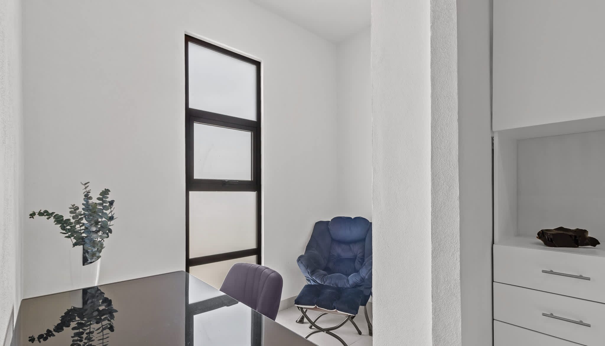 A modern room with a glass partition, a blue chair, a small plant, and a white cabinet.