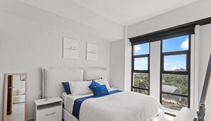 A bedroom with a large bed, a nightstand, a dresser, and a window with a view of greenery.