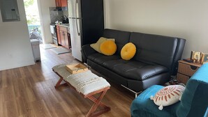 A modern living room with a black leather sofa, wooden coffee table, and a blue armchair. A kitchen is visible in the background with wooden cabinets and a stainless steel refrigerator. - Westside Magic 3bed 2 bath house in terrific westside of oahu!  (Nanakuli)