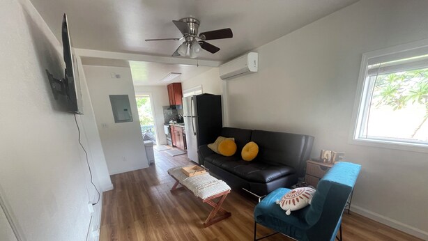 A living room with a black sofa, a blue armchair, wooden flooring, a ceiling fan, and a window with a view of greenery. - Westside Magic 3bed 2 bath house in terrific westside of oahu!  (Nanakuli)