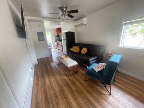A living room with a black sofa, a blue armchair, wooden flooring, a ceiling fan, and a window with a view of greenery.