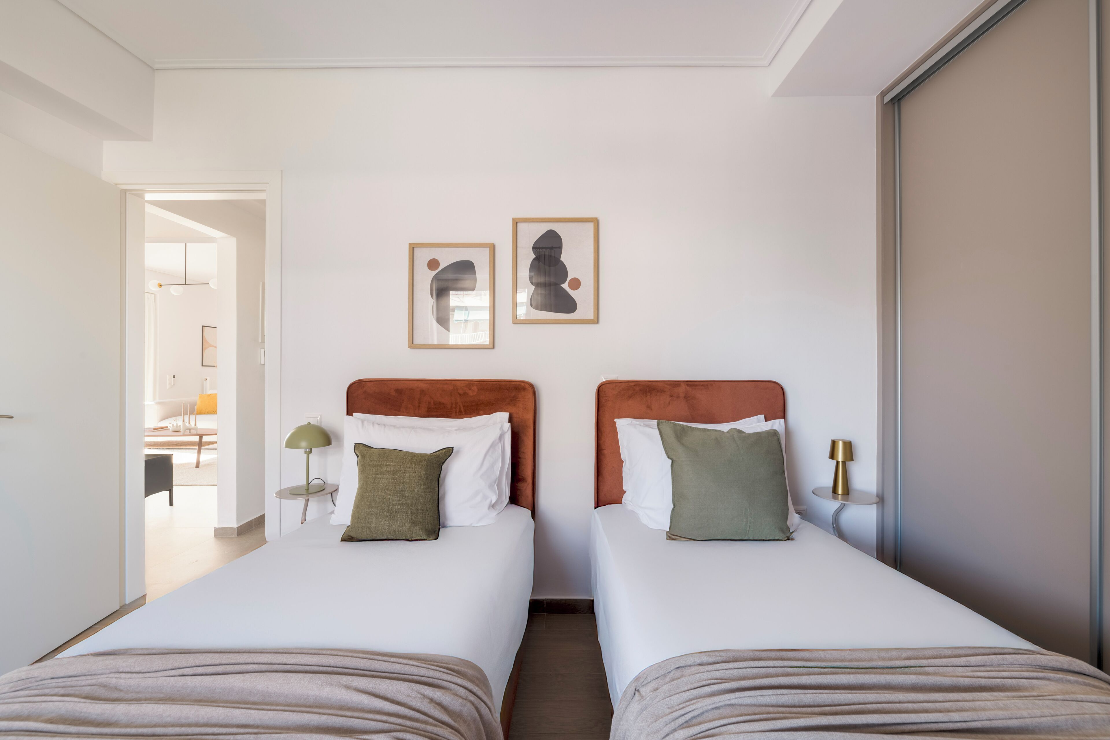 A hotel room with two beds, each with a wooden headboard, white bedding, and a green pillow. There are framed abstract art pieces on the wall, and a small table with a lamp beside the beds.