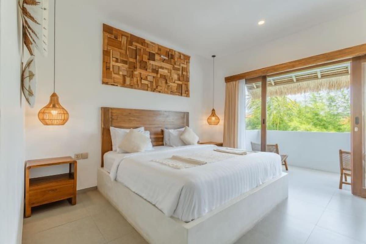 A spacious bedroom with a large bed, wooden headboard, and a view of greenery through a sliding glass door.