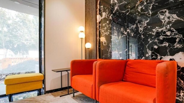 A modern room with a red sofa, a yellow ottoman, a small table, and a large window with a view of buildings. - Glitz Hotels DR Residency (Mumbai)