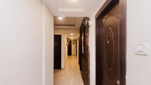 A hotel hallway with a dark wooden door on the right, a light-colored floor, and a ceiling light. - Pheonix By Glitz Hotels (Panvel)