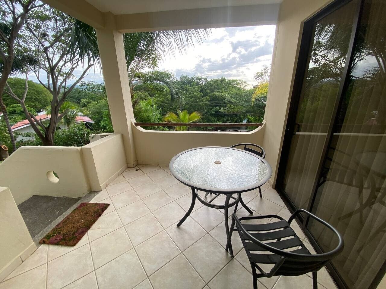 A balcony with a round table and chairs, a small rug, and a view of greenery.