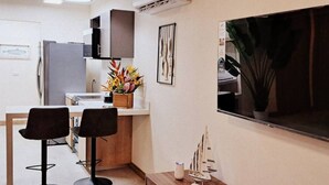 A compact kitchen with a refrigerator, two bar stools, a small table with a plant, and a mounted TV. - Brand New Condo - Heart of Coco! (Coco)