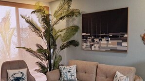 A living room with a beige sofa, two patterned pillows, a large potted plant, a framed abstract painting, and a chair with a crab design pillow. - Brand New Condo - Heart of Coco! (Coco)