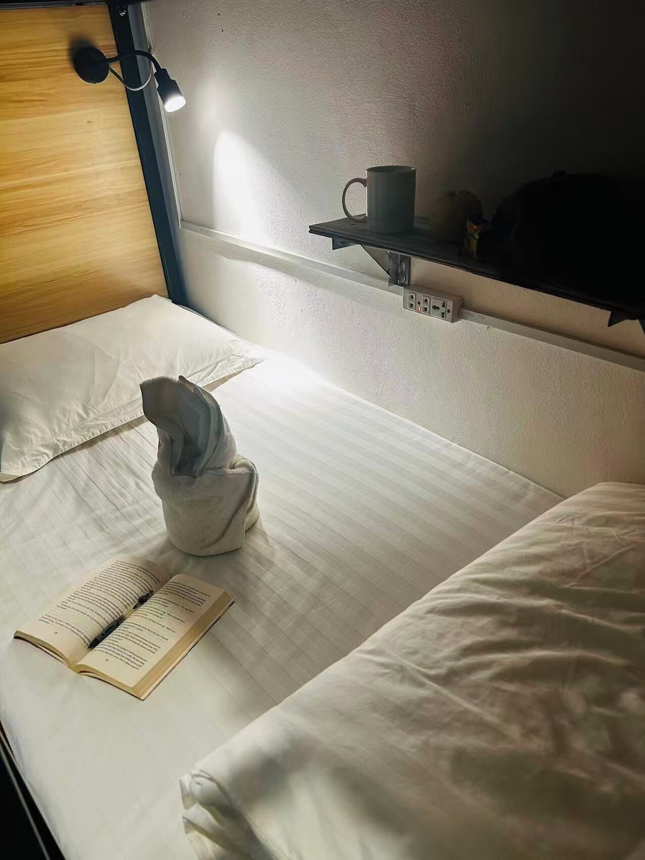 A single bed with a white mattress, a bedside lamp, a book, a towel, and a cup on a shelf.