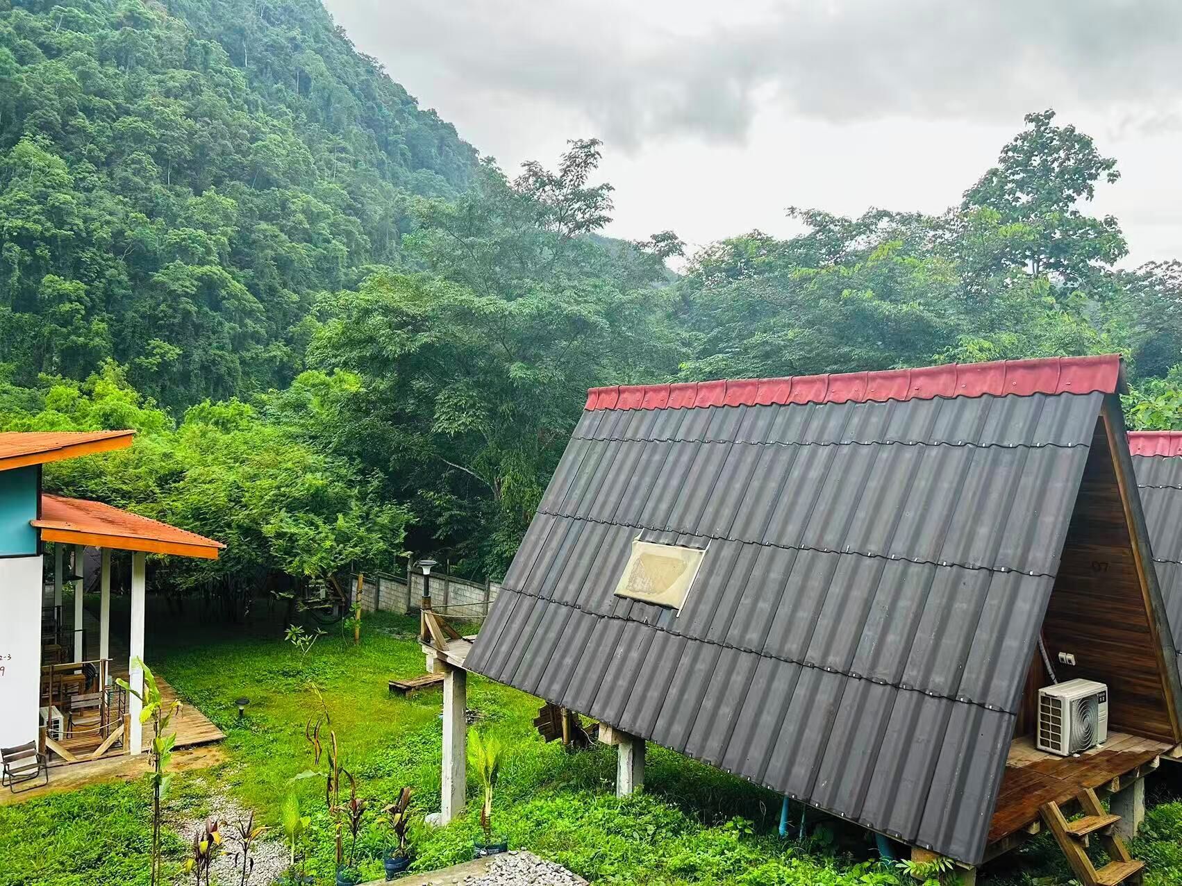 A rustic cabin with a sloped metal roof, surrounded by lush greenery and a mountain backdrop.