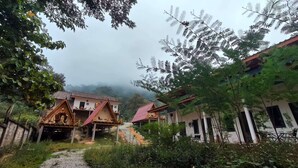 A rustic accommodation with multiple wooden structures, some elevated on stilts, surrounded by dense greenery and a foggy backdrop.