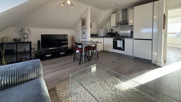 Eine moderne Küche mit weißen Schränken, ein Essbereich mit Glastisch und ein Wohnzimmer mit Sofa und Fernseher.