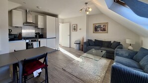 Eine moderne Küche mit weißen Schränken, einem Esstisch und einem roten Stuhl. Es gibt einen Wohnbereich mit einer Couch und einem Glastisch.
