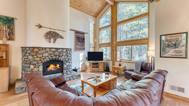 A cozy living room with a stone fireplace, leather sofas, and large windows offering a view of the forest. - Hansel by AvantStay | Pool Table, 5mins to Skiing (Truckee)