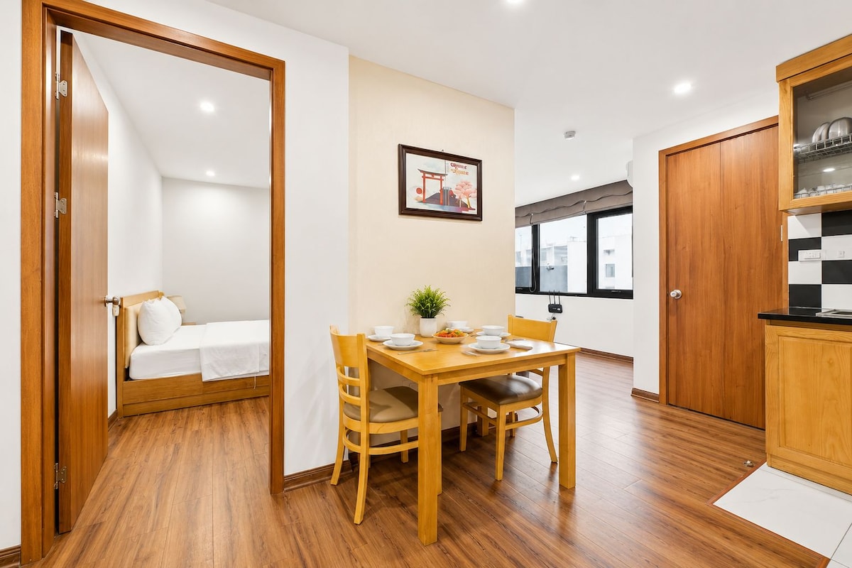 A compact room with a wooden floor, a dining table with chairs, a bed with a white mattress, a framed picture on the wall, and a kitchen area with cabinets.