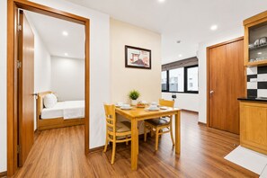 A compact room with a wooden floor, a dining table with chairs, a bed with a white mattress, a framed picture on the wall, and a kitchen area with cabinets. - Wendy apartment 39LL (Hanoi)