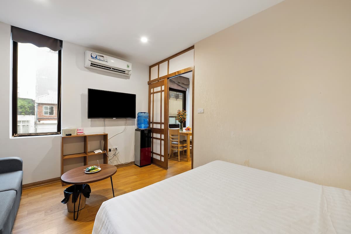 Une chambre d’hôtel comprenant un lit, un bureau avec une chaise, une télévision, un climatiseur et une fenêtre donnant sur des immeubles.