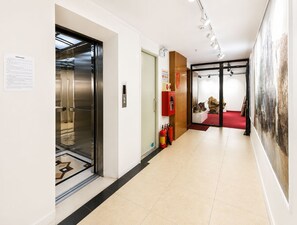 A hallway with an elevator, a fire extinguisher, and a red carpet leading to a room with a large abstract painting. - Wendy apartment 39LL (Hanoi)