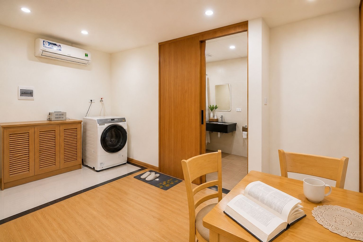 A compact room with a washing machine, a wooden cabinet, a dining table with chairs, and a small bathroom visible through an open door.