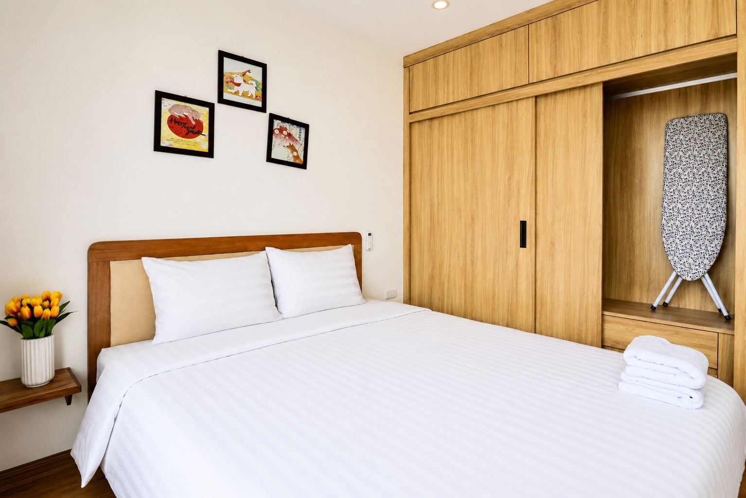 A neatly made bed with white linens, a wooden headboard, and a wall-mounted ironing board with a floral pattern.