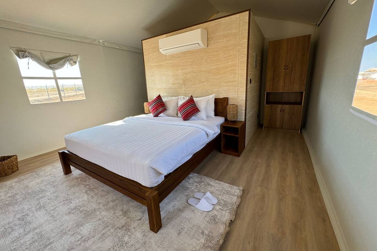 A bedroom with a wooden bed, white bedding, a small bedside table, a nightstand, a wooden wardrobe, and a window with a view of the landscape.