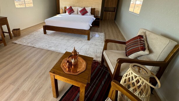A spacious bedroom with a large bed, a wooden nightstand, a wicker chair, and a small table with a decorative item. - Rahalla Luxury Stay (Bidiya)
