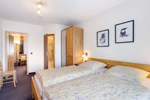 A hotel room with two beds, a wooden wardrobe, a chair, a desk, and two framed pictures on the wall.