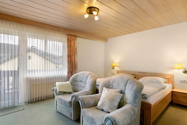 Ein Zimmer mit einem Bett, zwei Sesseln, einer Holzbalkendecke, einem Fenster mit transparenten Vorhängen und einer Wandlampe.