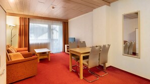 Habitación de hotel con alfombra roja, un sofá, una mesa de comedor con sillas, una lámpara y un ventanal con cortinas.