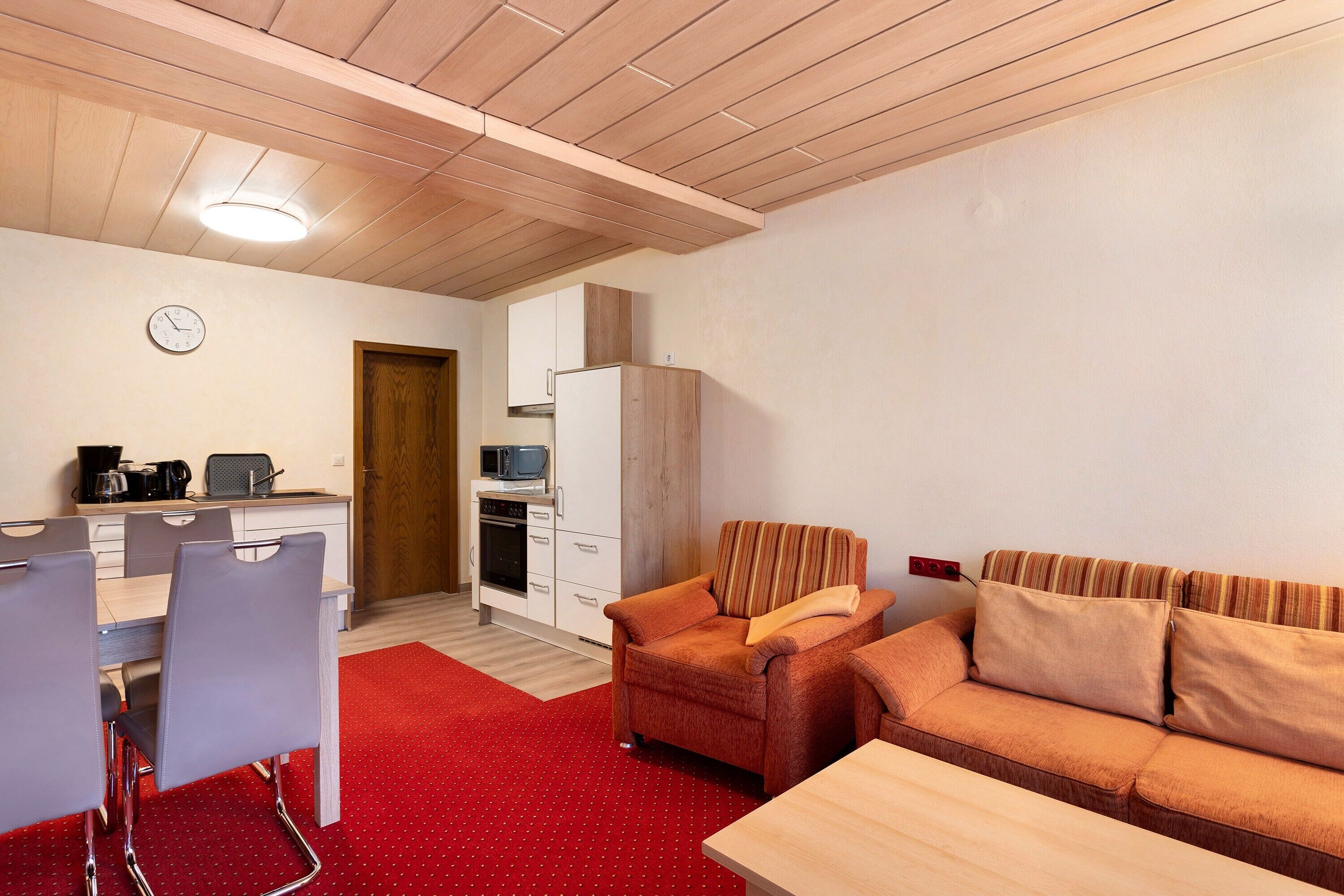 Una habitación compacta con kitchenette, una zona de comedor con sillas, un sofá y una mesa de madera.