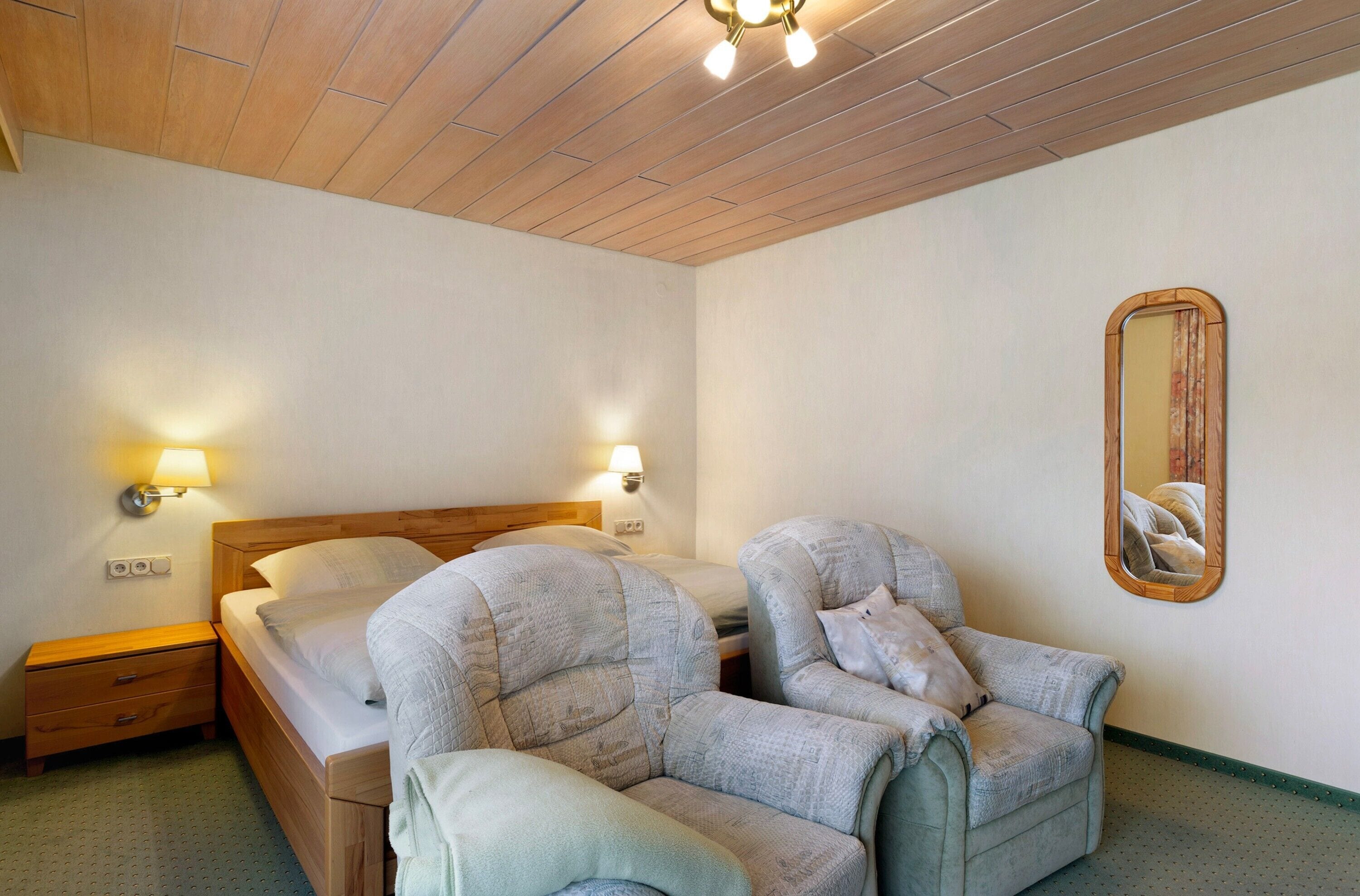 Habitació d'hotel amb sostre de fusta, un llit amb capçalera, dues butaques, una tauleta de nit i un mirall fixat a la paret.