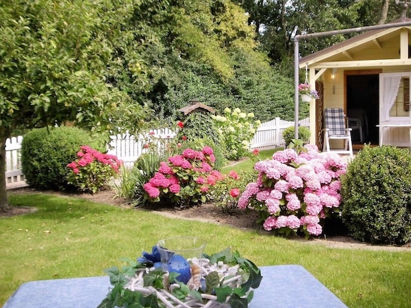 A garden with various plants and flowers, a white picket fence, and a building with a yellow facade.