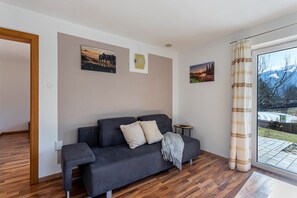 Een moderne woonkamer met een grijze bank, twee kussens, een klein bijzettafeltje en een groot raam met gordijnen.