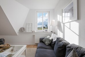 Una habitación con un sofá gris, un radiador blanco, una ventana con vistas al campo y un cuadro en la pared.