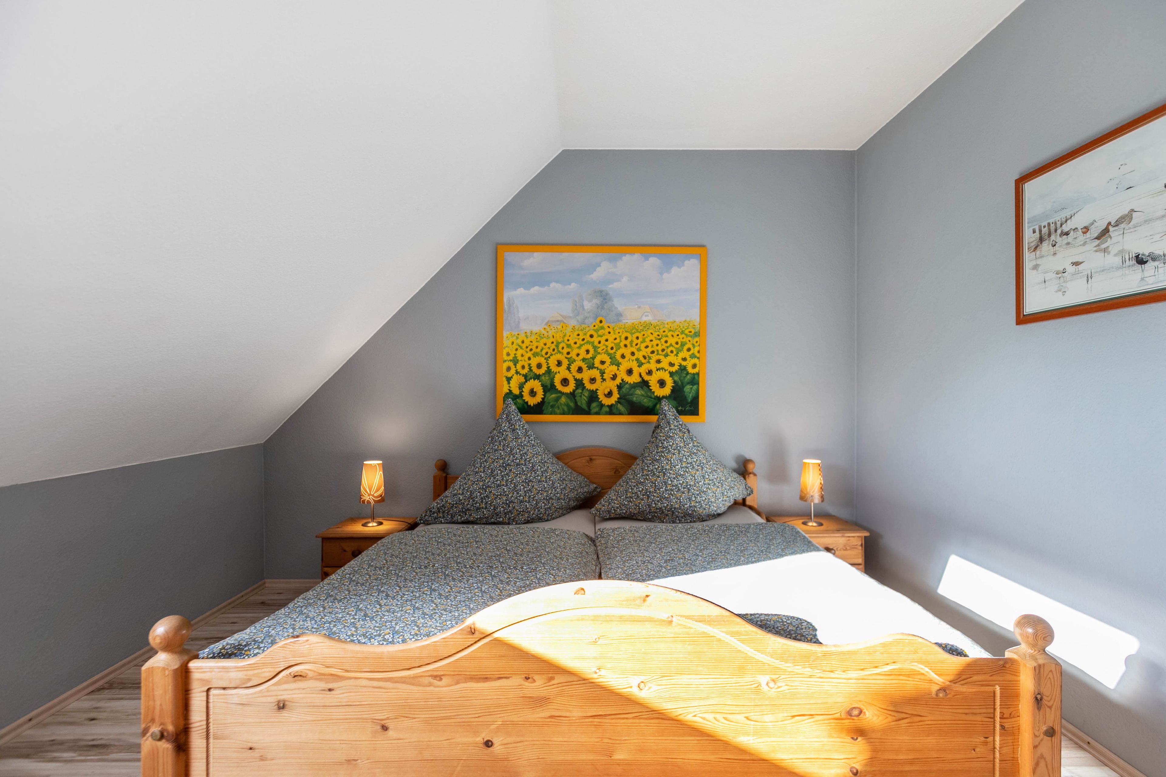 A bedroom with a sloped ceiling, a bed with a wooden headboard, two bedside tables with lamps, and a painting of sunflowers on the wall.