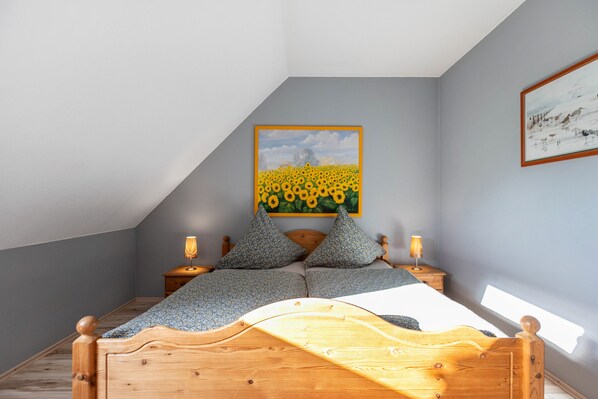 A bedroom with a sloped ceiling, a bed with a wooden headboard, two bedside tables with lamps, and a painting of sunflowers on the wall.