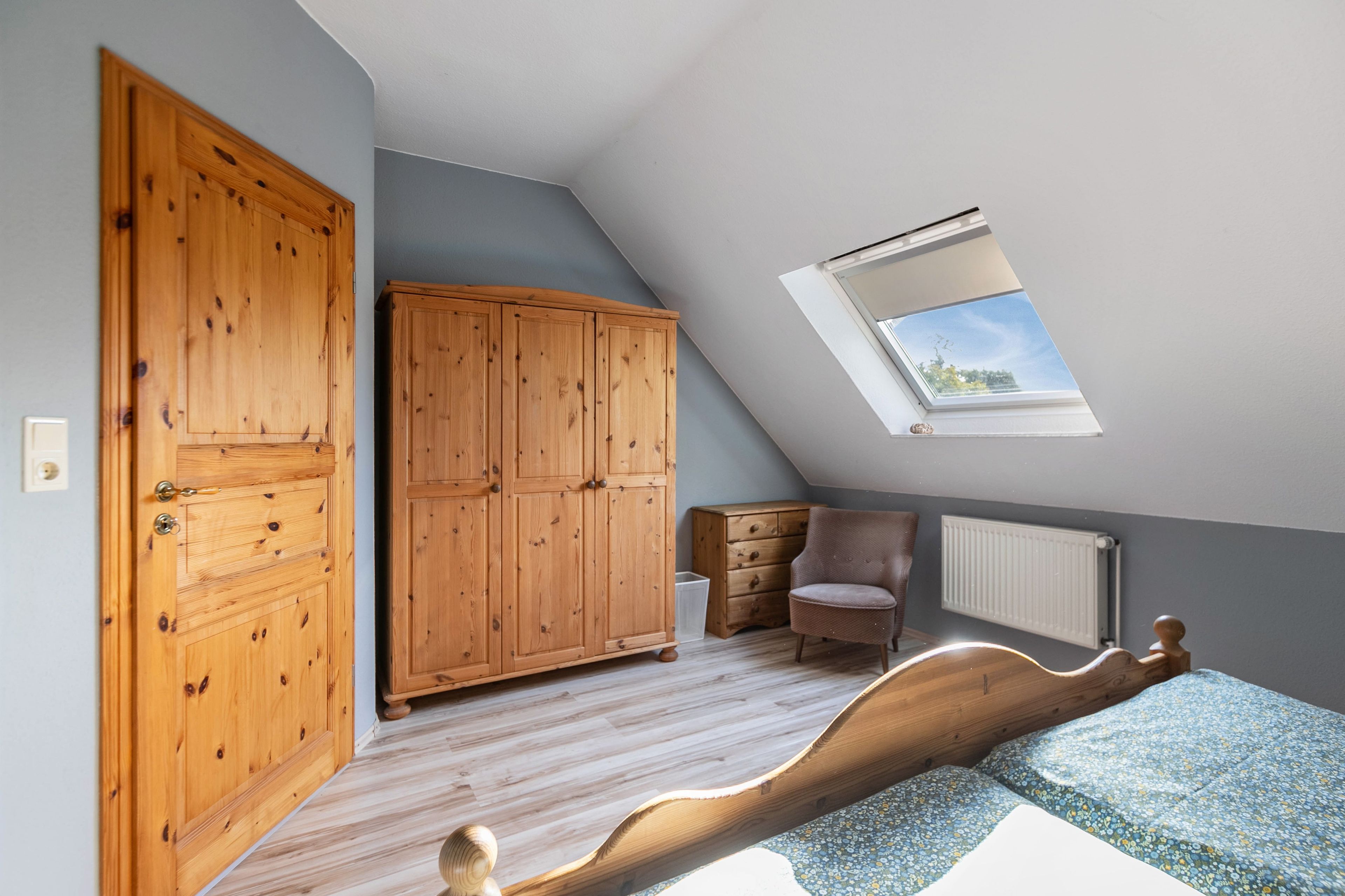 Ein Schlafzimmer mit schrägem Dach, einem Holzschrank, einem Sessel, einer Heizung und einem Oberlicht.