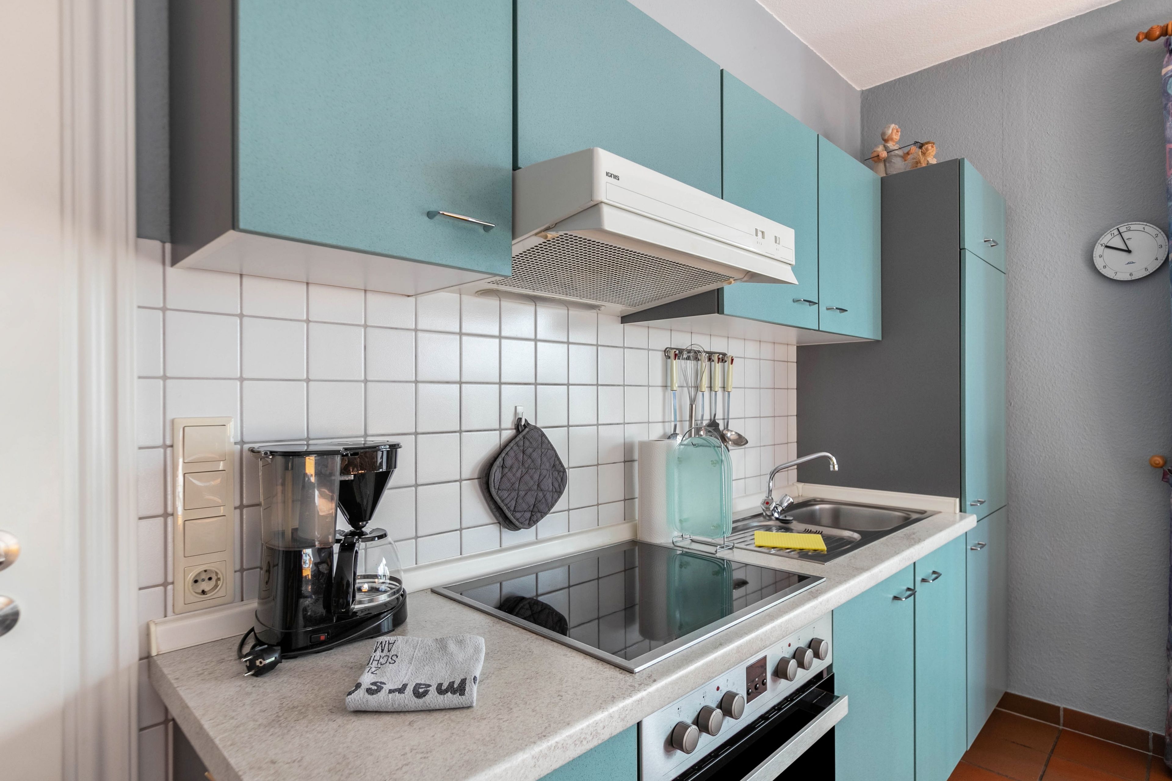 A kitchen with teal cabinets, a coffee maker, a kettle, and a clock on the wall.