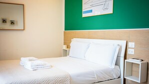 Una cama bien hecha con sábanas blancas y una toalla doblada, una mesita de noche con un control remoto, e interruptor de luz en la pared.