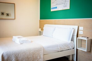 Una cama bien hecha con sábanas blancas y una toalla doblada, una mesita de noche con un control remoto, e interruptor de luz en la pared.