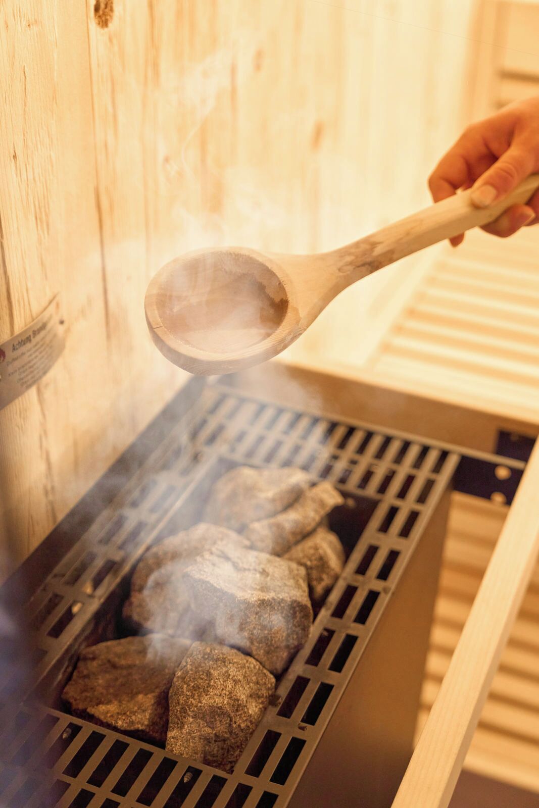 Una persona sosteniendo una cuchara de madera sobre una parrilla con piedras y humo.