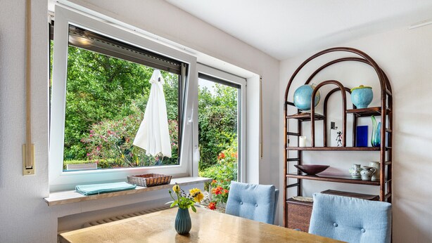 Een eetgedeelte met een houten tafel, stoelen, een vaas met bloemen, een raam met uitzicht op groen en een boekenkast met decoratieve objecten.