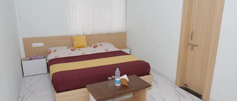 A bedroom with a bed, a wooden nightstand, a small table, a door, and a window with blinds.