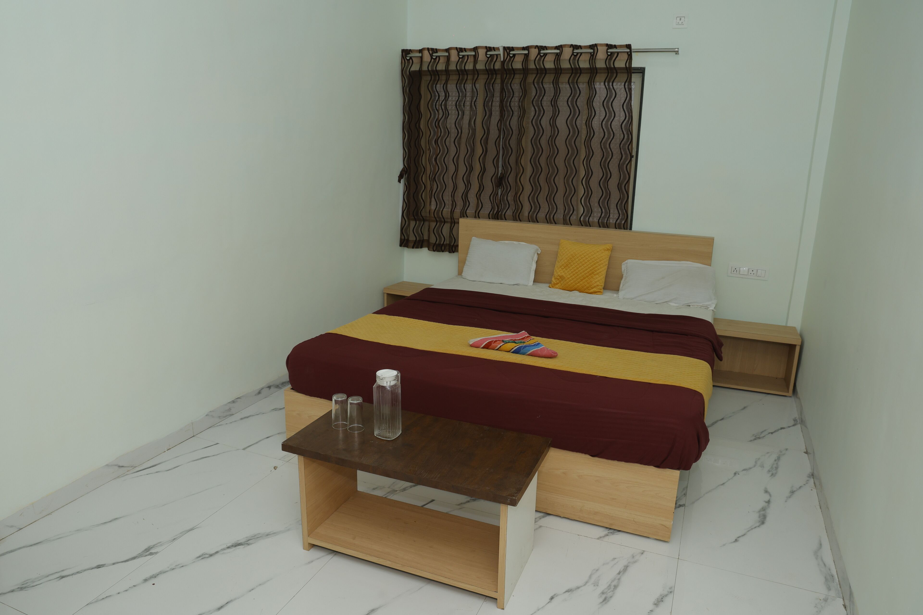 A bedroom with a bed, a small table with two glasses, a wooden nightstand, and a window with curtains.