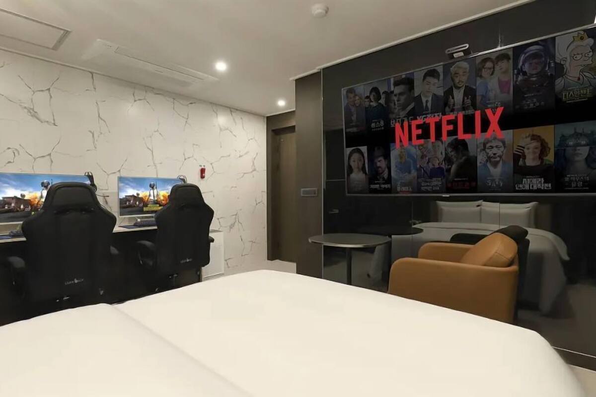 A hotel room with a large bed, two gaming chairs, a small table, a sofa, and a wall with a Netflix poster.