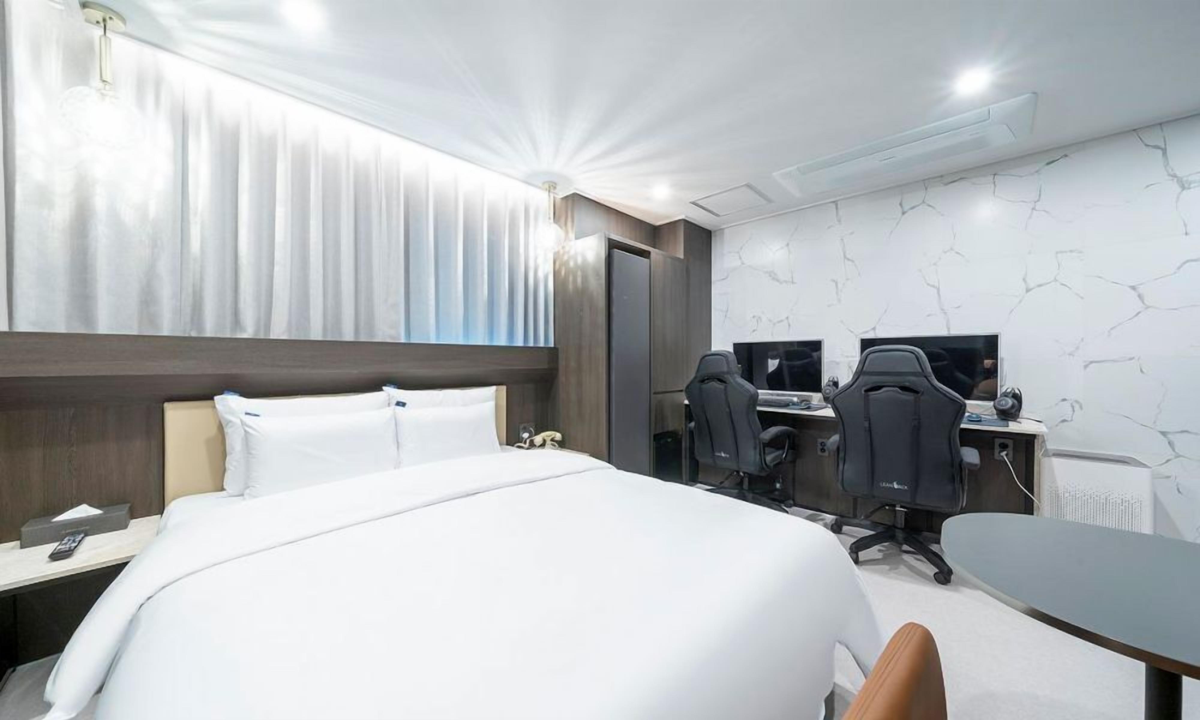 Quarto de hotel com uma cama grande, duas mesas com computadores e design moderno.
