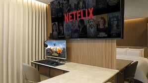 Habitación de hotel con un televisor de pared grande mostrando contenido de Netflix, un escritorio con computadora y una cama con cabecera.